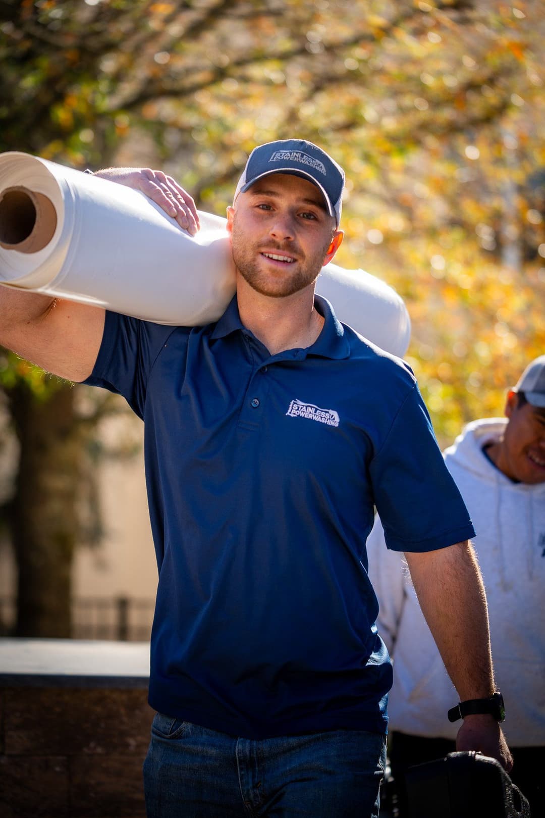 Man carrying large roll of material on shoulder, wearing a branded polo shirt and cap outdoors.