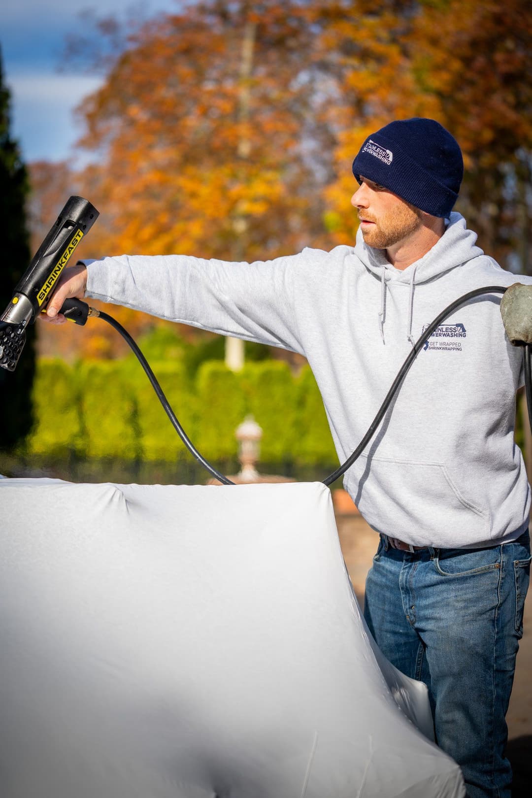 Man in hoodie using a heat gun to work on a white covering outdoors with fall foliage in background.
