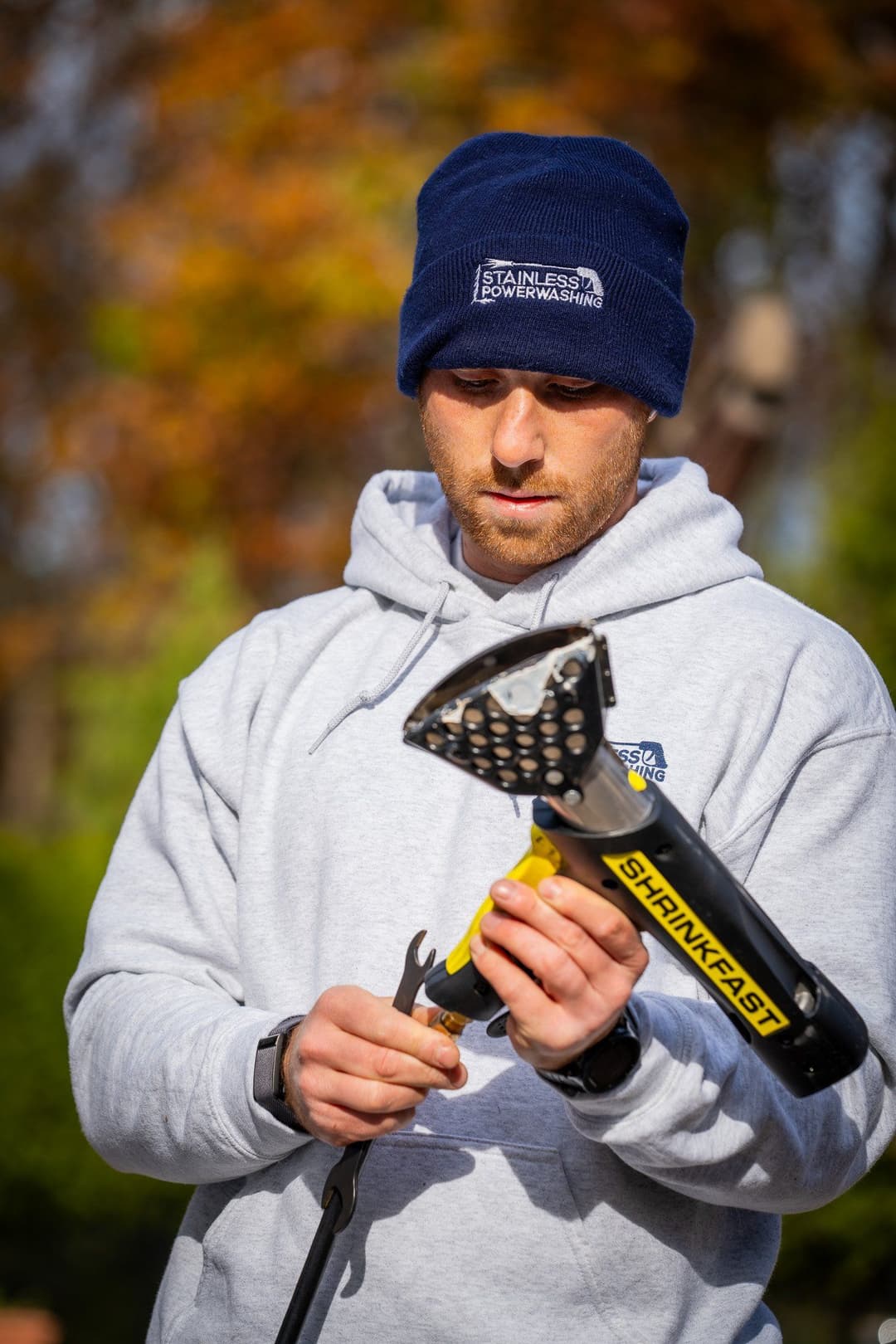 Man in gray hoodie inspecting a pressure washer attachment outdoors in autumn foliage.