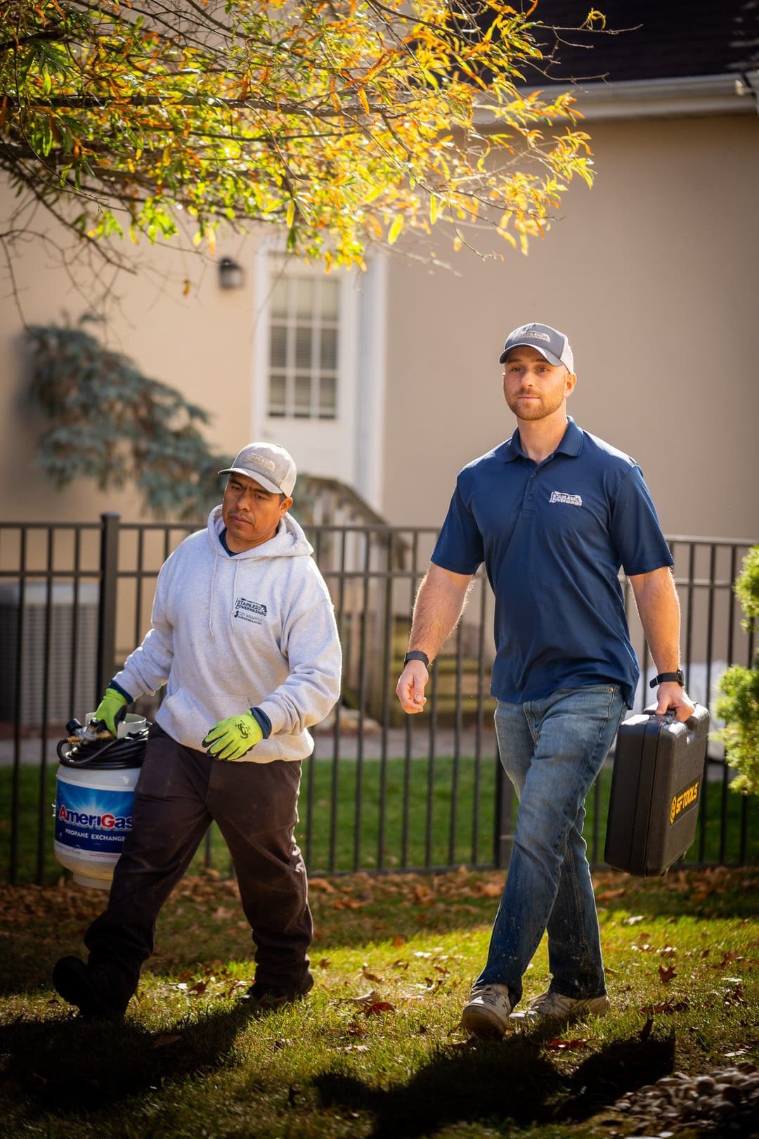 Two landscaping workers walking on a lawn, one carrying a bucket and the other a toolbox.