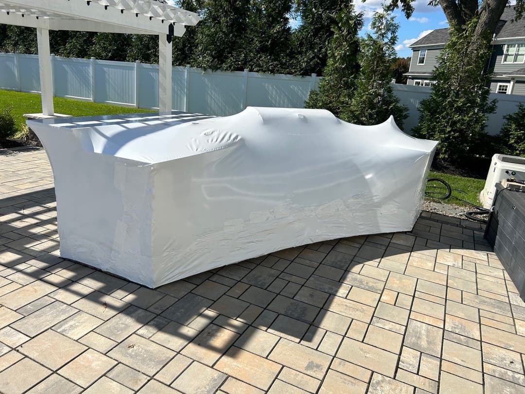 Covered outdoor hot tub with a white protective cover on a stone patio in a backyard setting.