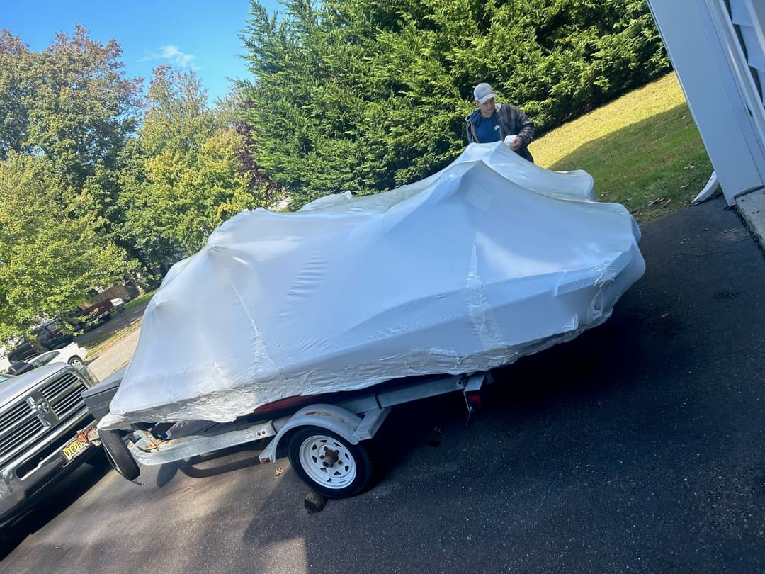 Person covering a car with a protective tarp in a driveway surrounded by trees.