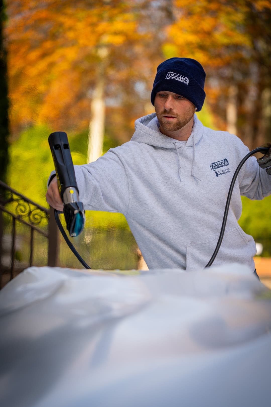 Man in a gray hoodie and beanie using a heat gun on a white surface outdoors.