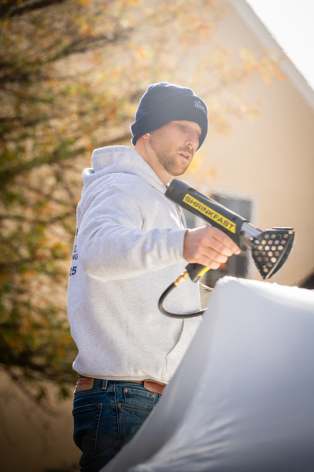 Man using a heat gun to shrink wrap an object outdoors, wearing a gray hoodie and beanie.