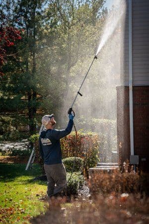 Man using a pressure washer on a house exterior with trees in the background.
