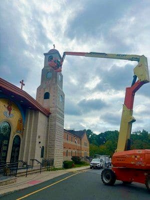 Worker on a lift performing maintenance on a church steeple under a cloudy sky.
