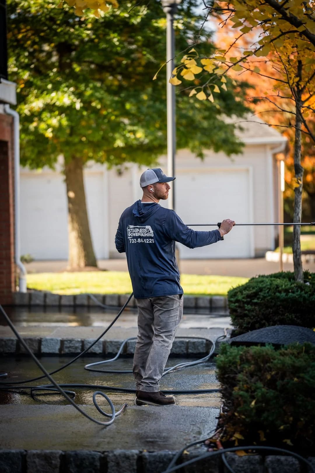 Man power washing a driveway with hoses and greenery in the background during autumn.
