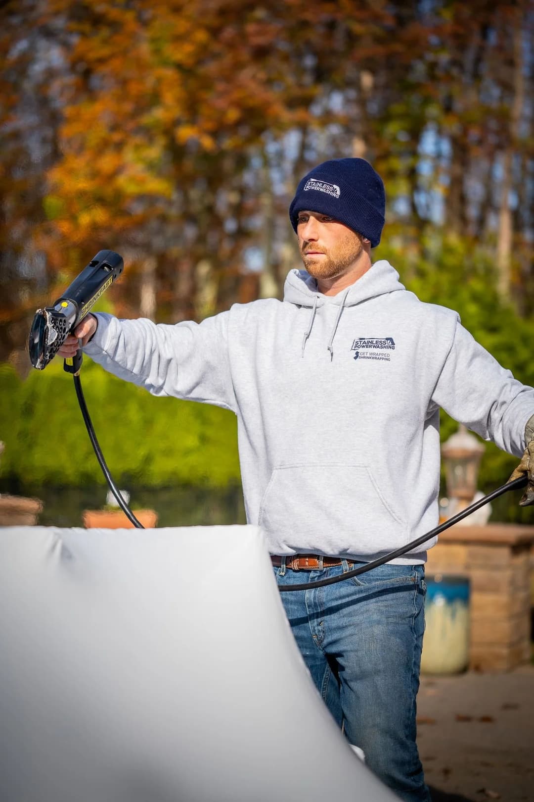 Man wearing a hoodie and beanie operates a spray gun outdoors for coating a surface.