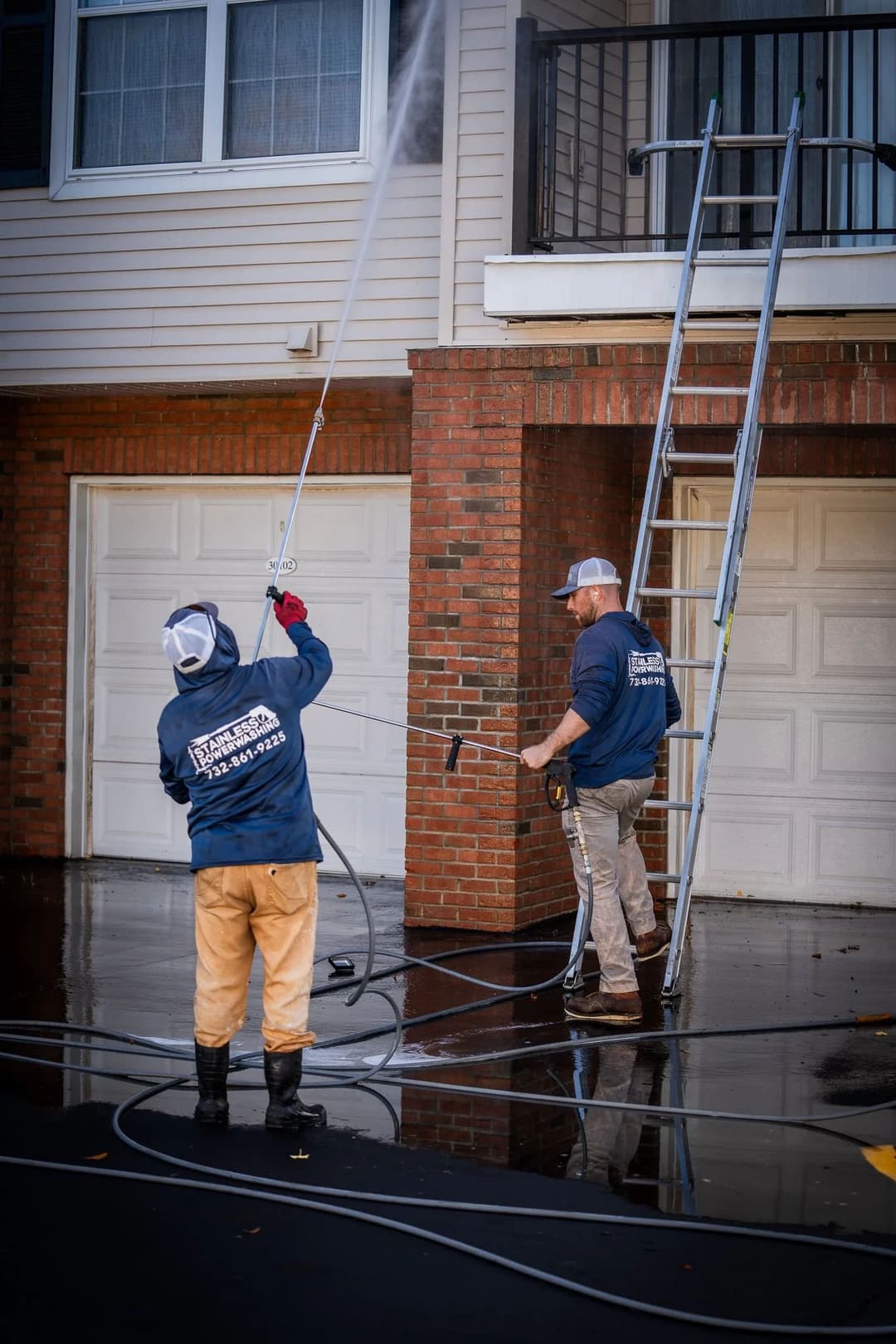 Two workers pressure washing a building exterior with ladders and equipment.