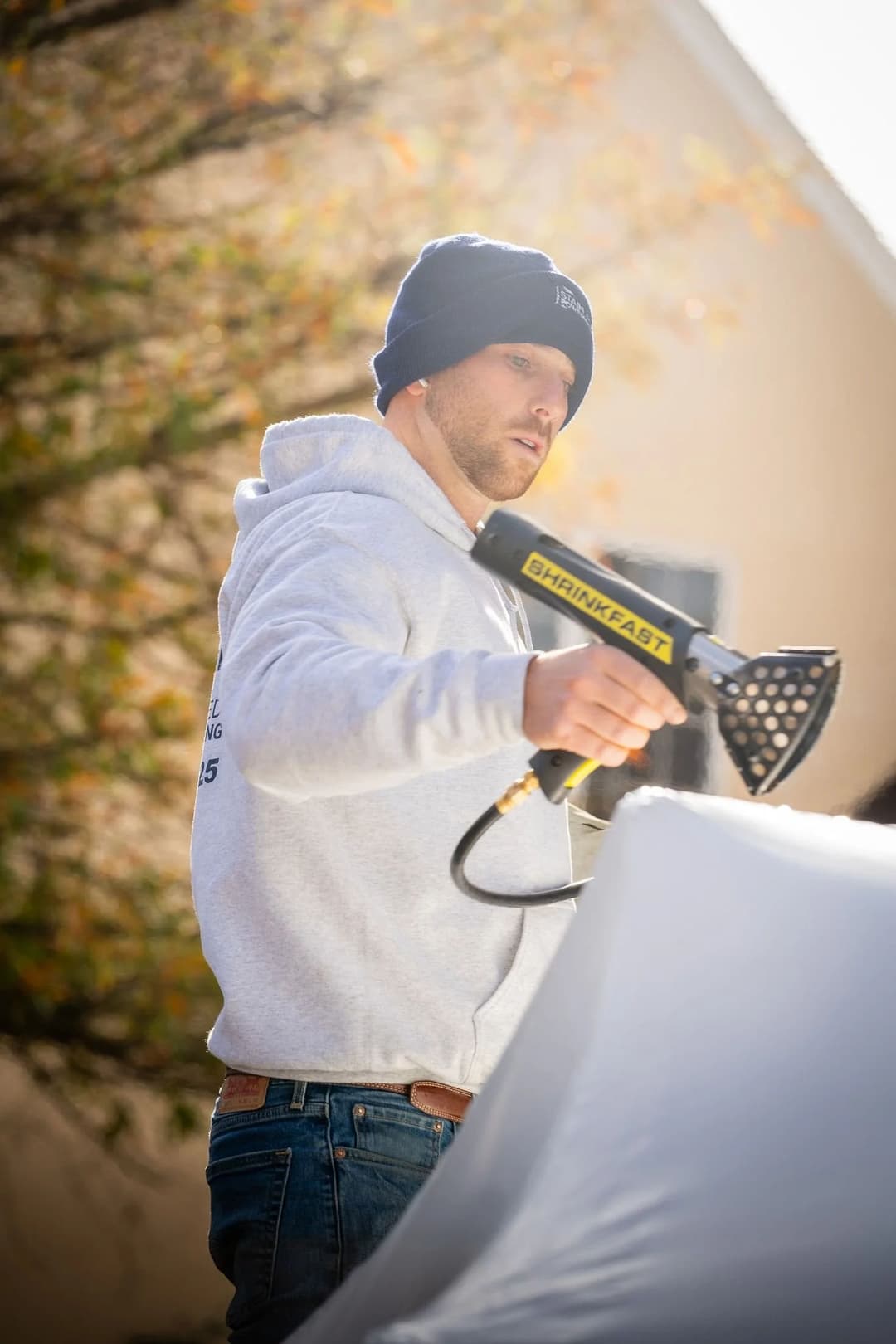 Man using a heat shrink gun on a white covering outdoors, wearing a hoodie and beanie.