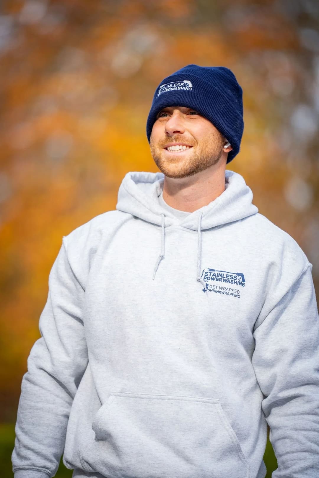 Man smiling outdoors in gray hoodie and blue beanie, with colorful autumn background.